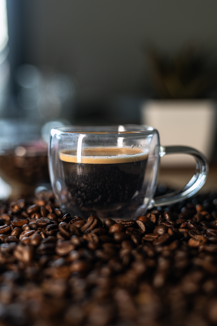 Glass cup of freshly brewed coffee surrounded by aromatic coffee beans on a rustic surface.
