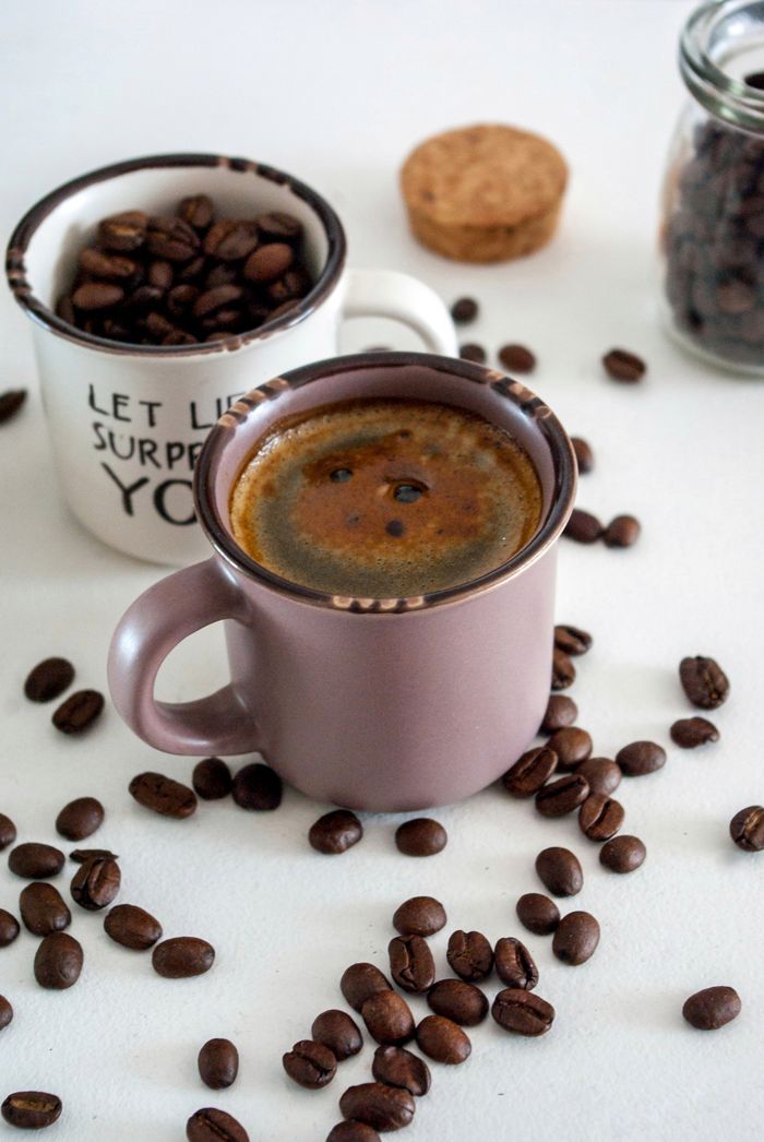 Close-up of a cup of espresso surrounded by scattered coffee beans on a white surface.