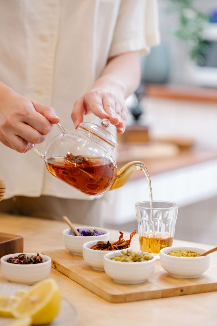 A person pouring herbal tea into a glass with various ingredients on a wooden board.